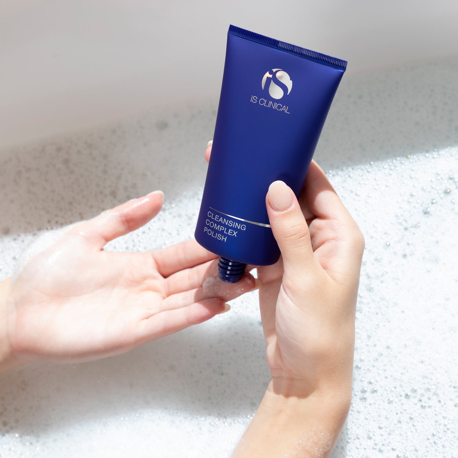 Hand holding IS CLINICAL Cleansing Complex Polish tube over foam in a relaxing bath setting.